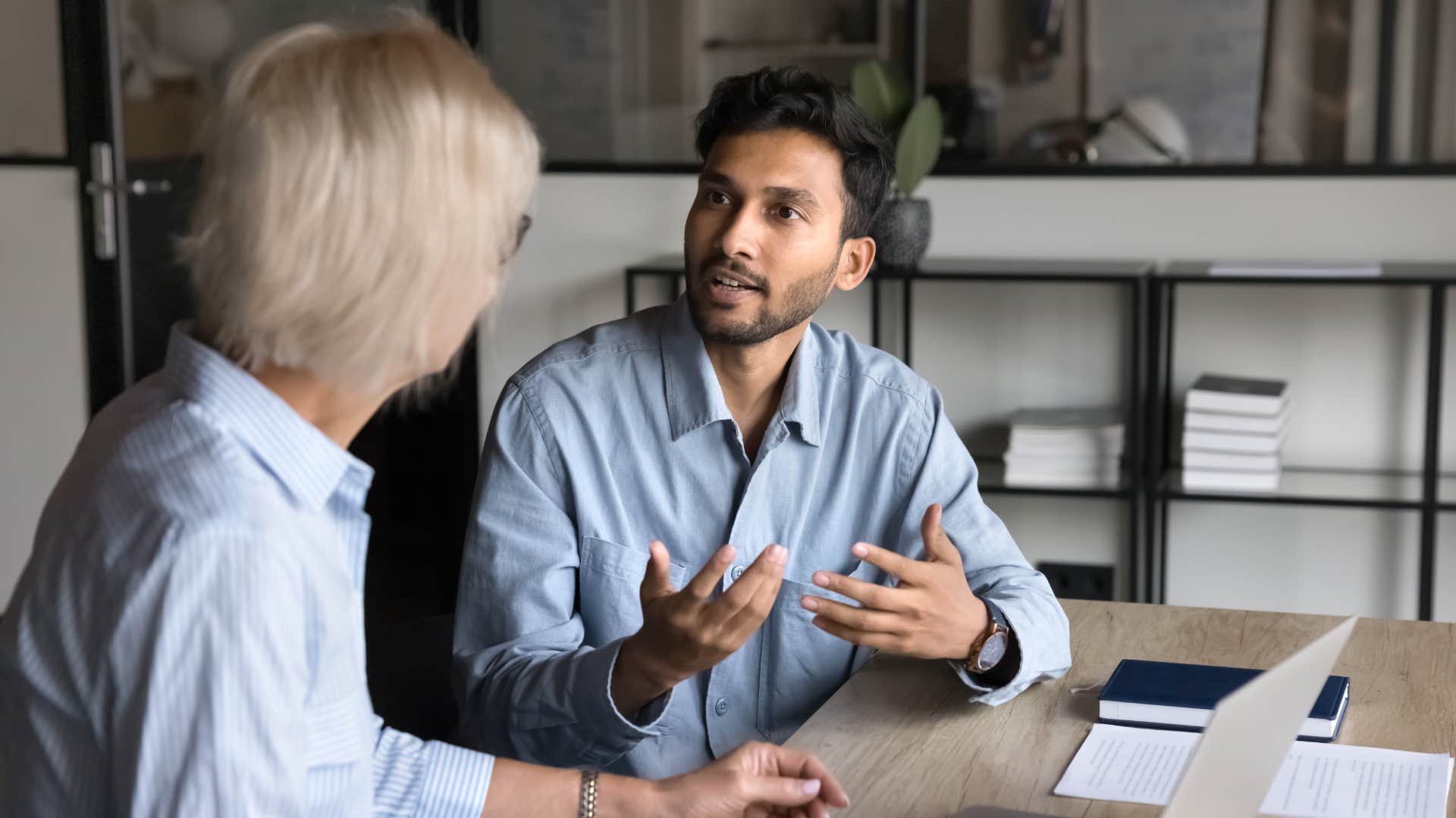 man in blue shirt asking woman is it possible this event is going to go overtime while they sit down in front of laptop